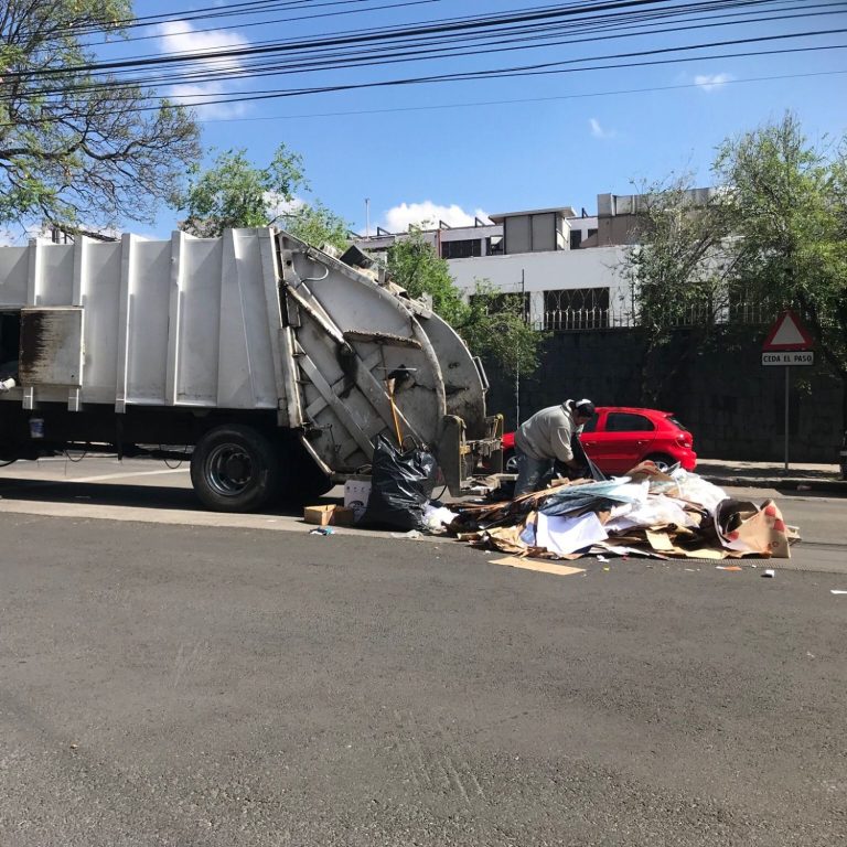 Camión de basura recogiendo desechos en la calle junto a un coche rojo estacionado.